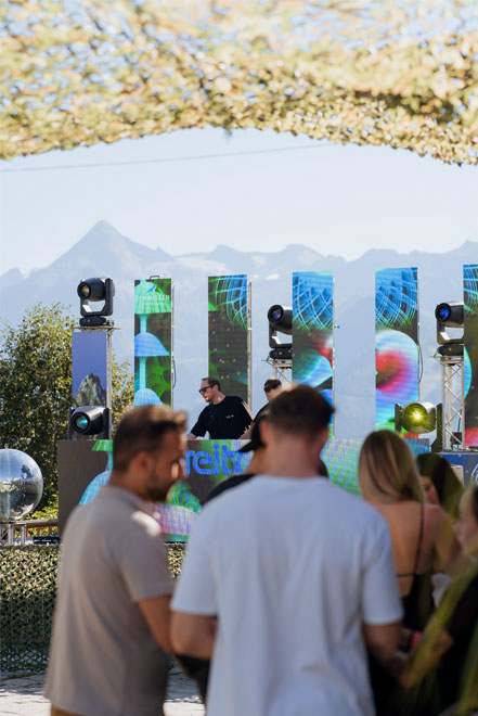 DJ performt tagsüber vor LED-Wänden und feiernder Crowd mit Blick auf die Berge.