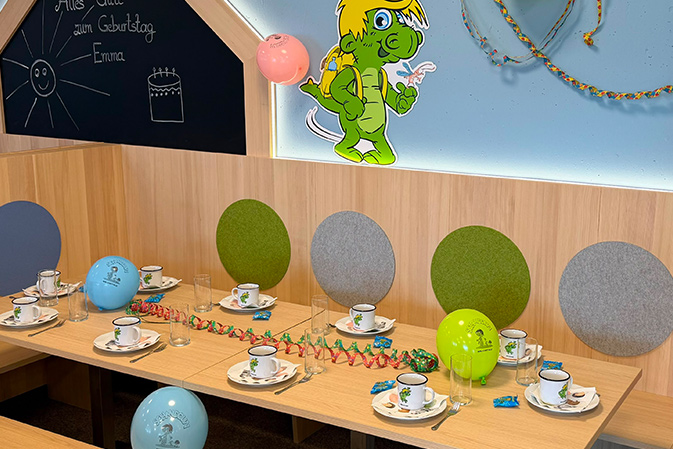 Geburtstagsdekoration mit bunten Luftballons, einem Cupcake mit brennender Kerze, einem Geschenk mit buntem Papier und einer Tafel mit der Aufschrift 'Happy Birthday Emma' in einem Raum mit Holztischen und gepolsterten Wandpaneelen.