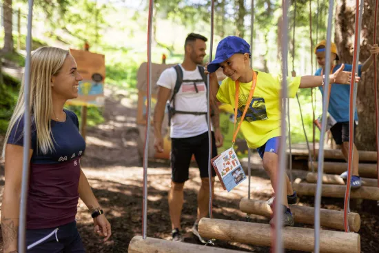 Kind mit blauer Kappe balanciert auf hängenden Holzstämmen im Kletterparcours, zwei Erwachsene beobachten im Wald